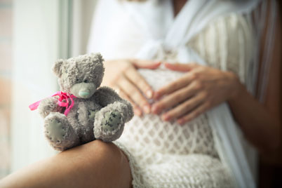 sitzende Schwangere mit ihren Händen auf dem Babybauch udn grauem Teddybär, der eine pinkfarbenes Halsband trägt,  auf dem Knie