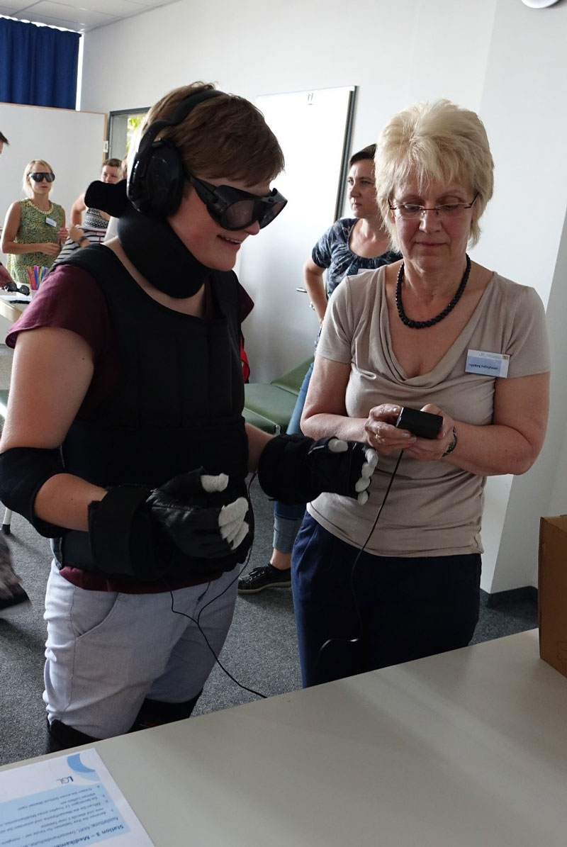 Eine Mitarbeiterin der Bayerischen Gesundheitsagentur testet gemeinsam mit einer Stipendiatin die Tremorhandschuhe. Die Teilnehmerinnen und Teilnehmer können dadurch nachempfinden, welche Alltagsprobleme durch das Alterszittern verursacht werden.