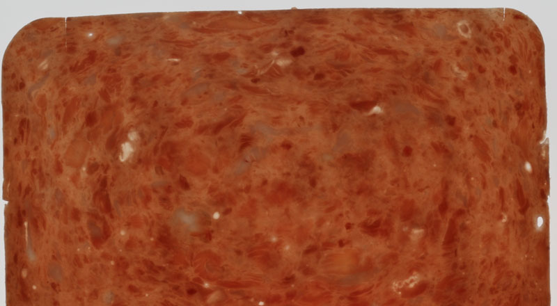Foto eines Schinkenimitats, bei der in einer homogenen Masse eingelagerte Fleisch-, Fett- und Bindegewebsstücke zu erkennen sind. Diese Stückchen reiskorn- bis etwa haselnussgroß.