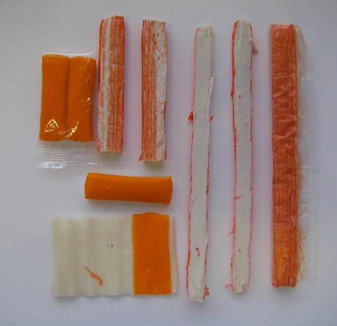 Das Bild zeigt unterschiedlich große Surimi-Sticks (weißfleischige Stangen aus mehreren aufgerollten Schichten, mit orangefarbenem  Paprikaextrakt gefärbt