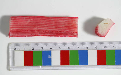 Das Foto zeigt von oben ein etwa 7 cm langes, 1,5 cm breites Stück Surimi mit roter Oberfläche. Daneben befindet sich ein kleines abgeschnittenes Stück, bei dem der Querschnitt sichtbar ist.