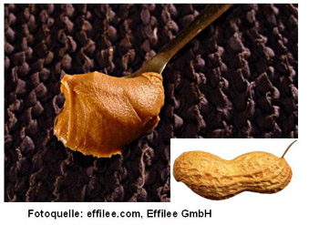 Es ist eine Erdnuss in der Schale neben einem Löffel mit dunkelbrauner Erdnusscreme dargestellt.