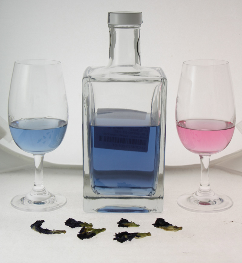 In der Abbildung ist eine geöffnete Flasche Gin zu sehen, der mit einem Extrakt aus der Blauen Klitorie hergestellt ist. Der blaue Gin ist auch in einem Glas links von der Flasche enthalten. Rechts von der Flasche befindet sich ein Glas mit dem rosafarbenen Gin, wie er nach Zugabe einer saueren Flüssigkeit entsteht. Im Vordergrund sind getrocknete Blüten der Blauen Klitorie zu sehen