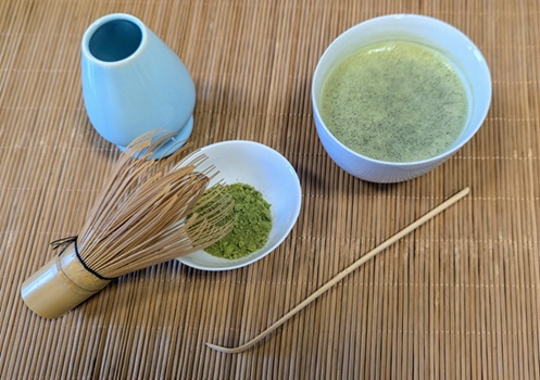 Auf dem Foto sind auf der linken Seite eine Schale mit Matcha-Teepulver, ein Bambusbesen und ein Matcha-Löffel und auf der rechten Seite ein zubereiteter Matcha-Tee abgebildet. 