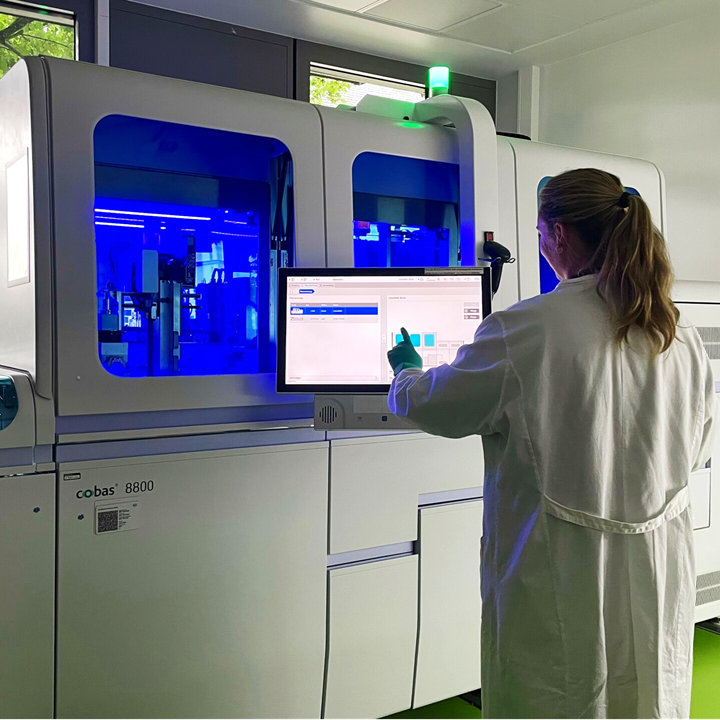 Wissenschaftlerin in Laborkittel vor Analysegerät mit blauem Licht. 
